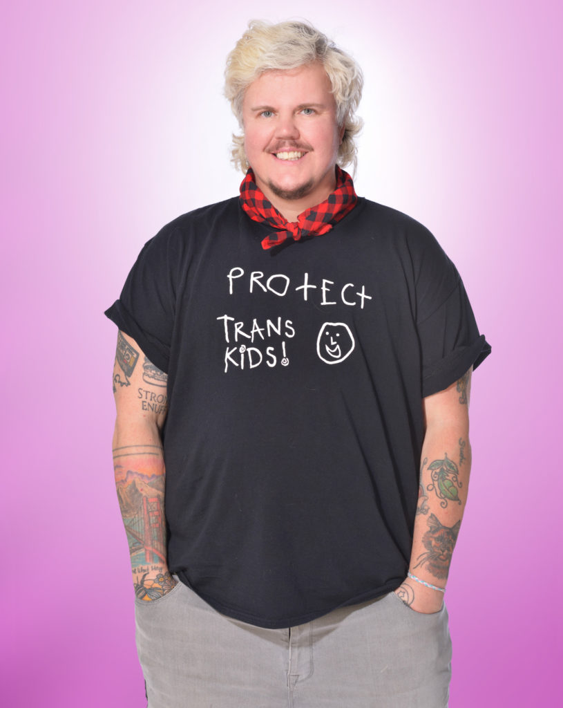 Kaleb, a white trans person with blond hair and brown facial hair stands with his hands in his pockets and is smiling. He is wearing grey jeans, a black tshirt that says "protect trans kids" with a smiley face, and a red and black bandana tied around his neck. He has colourful tattoos on his forearms. 